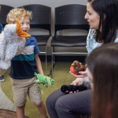 A librarian interacts with a little boy with curly blonde  hair. He is holding a duck puppet. Both are smiling.