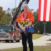 Former Mayor Gary Waters wears a Hawaiian shirt and stands in front of a fire engine with a large American flag.