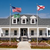 Exterior of Pelham City Hall. A white brick building with white columns and dormer windows. Steps lead up to a covered front porch.Two flags fly out front.