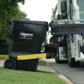 An Amwaste garbage truck reaches out with a mechanical arm to empty a trash can.