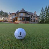 A golf ball with the Ballantrae logo sits on a green with the clubhouse in the background.
