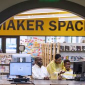 A family, including a man, woman, and a boy, look at a computer in the library.
