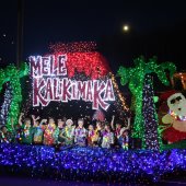 Children wearing Hawaiian shirts ride on a brightly colored float with the words, "Mele Kalikimaka" on the float.